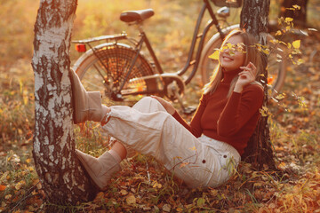 Happy active young woman sitting near vintage bicycle bike in autumn park at sunset. European...