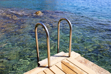 Metal ladder to the sea on wooden pier, view to transparent water and stones on a bottom. Beach vacation and swimming concept