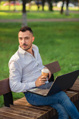 Man looking at the camera in the park with a laptop, drinking coffee. A young man on a background of green trees, a hot sunny summer day. Warm soft light, close-up.