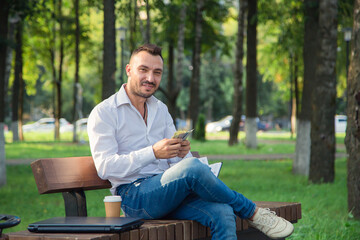 A smiling man is sitting on a bench in the park, counting the profit. A young man on a background of green trees, a hot sunny summer day. Warm soft light, close-up.