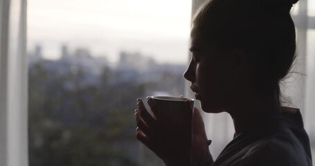 Young woman drinking coffee or tea while looking out window at home. Female having a quiet moment, enjoying the view and a calm, relaxed evening. Lady cooling a warm drink, relaxing after a long day - Powered by Adobe