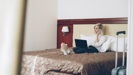 Smiling blonde businesswoman in white shirt using on laptop and talking at mobile phone while lying on bed in hotel room. Business, travel and people concept