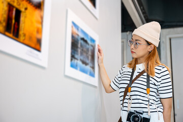 Asian woman hold camera at art gallery collection in front framed paintings pictures on wall and...
