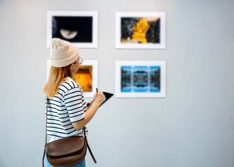 Young person at photo frame write digital book leaning against at show exhibit artwork gallery, Asian woman holding tablet at art gallery collection in front framed paintings looking pictures on wall - Powered by Adobe