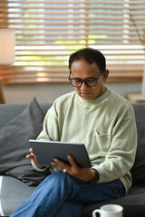 Happy mature man reading electronic book on digital tablet while relaxing in living room