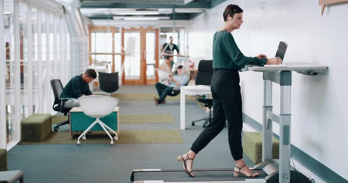 Female entrepreneur multitasking while walking on treadmill and doing research on laptop while listening to music in modern office. Fitness and work ambitions while exercising in coworking workplace