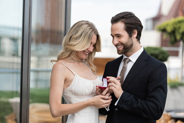 Cheerful man in suit holding box with engagement ring near girlfriend on terrace.