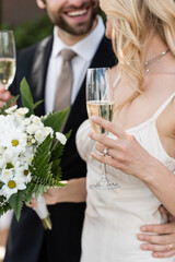 Cropped view of blonde bride holding bouquet and champagne near groom outdoors.