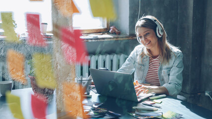 Creative young businesswoman is listening to music in headphones dancing while working at desk with laptop in modern office indoors. Glass with colored stickers in foreground.