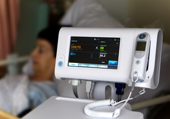 A heart/blood pressure monitor in a hospital ward with a blurred female patient in bed in the background