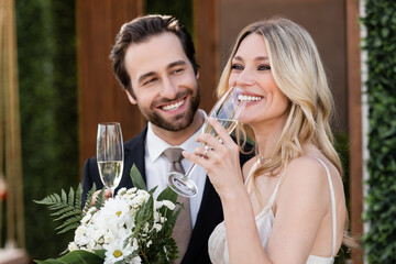 Smiling bride holding glass of champagne near blurred groom and flowers outdoors.