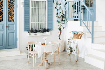 Exterior of a mediterranean style white house with a blue door and window, flowering tree and a romantic seating area with a vintage white bicycle with flowers in a basket. House in Santorini