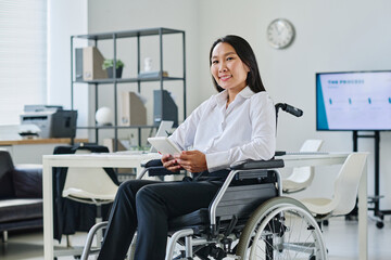 Portrait of Asian businesswoman sitting on wheelchair and smiling at camera while using digital...