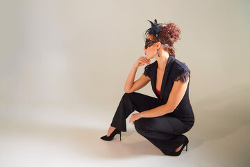 Portrait of beautiful slim young woman with short brunette hair in an elegant black jumpsuit and a mysterious mask in studio with white background. Girl model posing during photo shoot