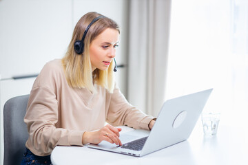 Stylish young woman video conference via online chat, wearing a wireless headset, sitting at home in front of an open laptop. Electronic gadgets and communication concept