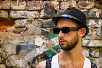 Portrait stylish bearded young man in sunglasses with suspenders and hat at brick wall background. Face of chic posh confident guy of sexy look outdoors. Concept of style, fashion, beauty. Copy space