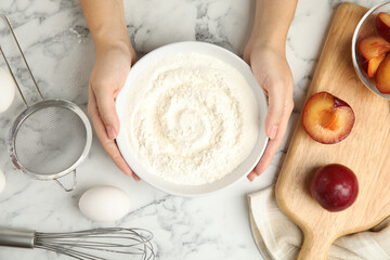 Fototapeta premium Woman holding bowl with flour at white marble table, top view. Cooking of delicious plum cake