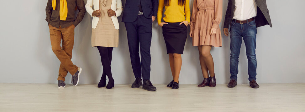 People in smart casual clothes by office wall. Row of company employees in pants, skirts, jackets and jumpers of brown, yellow, dark blue, beige color. Low section legs. Work dress code concept header