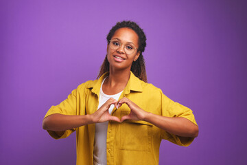 Young attractive African American woman show heart gesture symbolizing not indifference to needy and charitable activities or desire to be volunteer stands in purple studio. Mercy, help