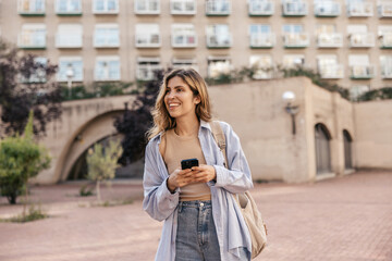 Nice young caucasian woman holding smartphone in her hands spending leisure time outdoors. Blonde wears tank top, shirt, jeans and backpack. Concept lifestyle, devices