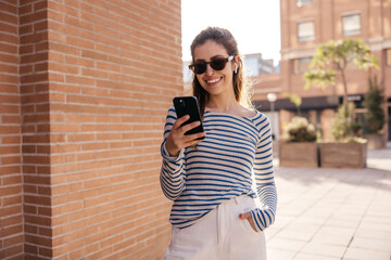 Positive young caucasian woman using sms chat on smartphone spending leisure time outdoors. Brown-haired model wears sunglasses, jacket and pants. Technology concept