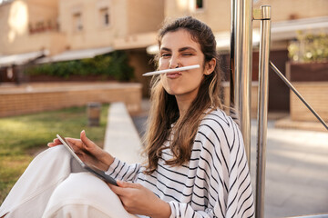 Funny young caucasian girl grimaces holding stylus in place of her mustache relaxing outdoors....