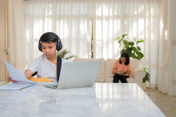 Asian boy playing guitar and watching online course on laptop while practicing for learning music or musical instrument online at home. Boy students study online with video call teachers play guitar.