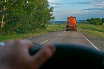 view of a fuel tanker with an orange tank carrying flammable cargo on a country road against the background of a driver who is driving behind a truck