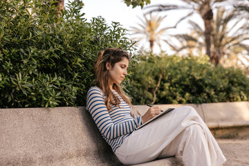 Pretty young caucasian woman makes notes in notebook sitting outdoors. Brown-haired girl wears casual clothes spring. Leisure concept