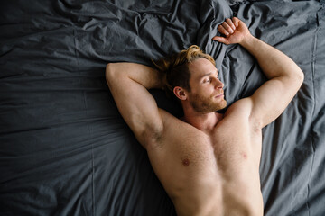 Young bearded shirtless man smiling and lying in bed at home