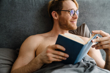 Young shirtless man wearing eyeglasses reading book in bed at home