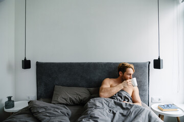 Fototapeta premium Young bearded shirtless man drinking coffee while lying in bed at home