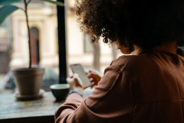 Young black woman using cellphone and drinking coffee in cafe indoors