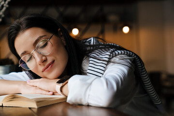 White brunette woman in eyeglasses reading book while sitting at cafe