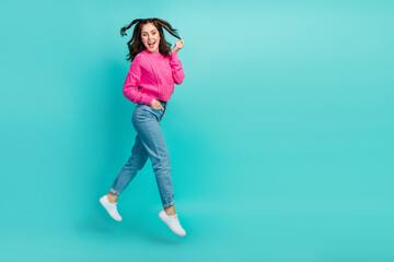 Fototapeta na wymiar Full size photo of cute girl with wavy hairdo pink pullover jeans sneakers hand in pocket touch hair isolated on teal color background