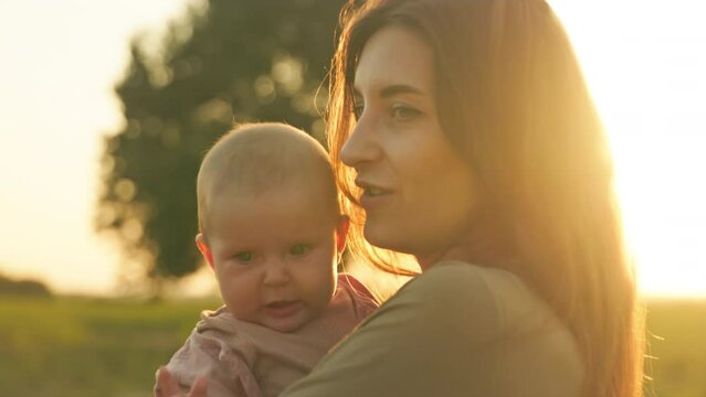 Close-up view of beautiful Caucasian woman in casual clothes holding crying little baby girl in hands, kissing lovely daughter. Blurred background of old green thee. Field. Amazing sunset. Outdoor