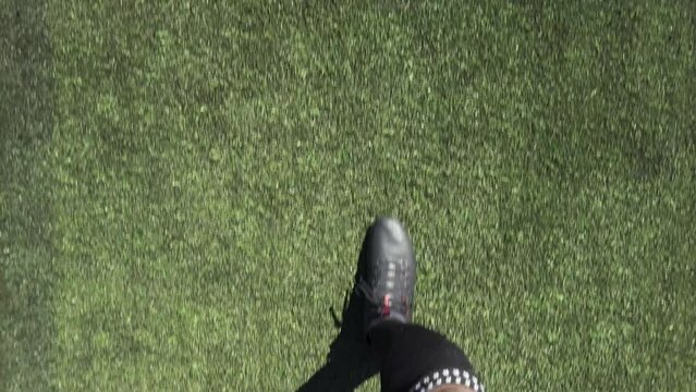 POV shot looking down at a football player's boots as he walks across an artificial soccer pitch on a sunny day