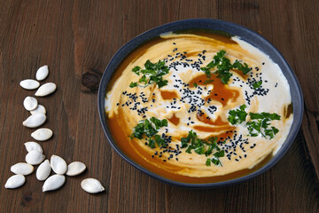 Bowl of creamy pumpkin soup with cream, black cumin seeds, herbs and cranberries on a wooden table