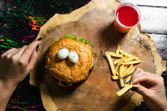 Top View Of An Order Of Hamburger With Fries And Red Fruit Juice, Served On A Wooden Tray In The Shape Of A Tree Trunk. Latina Girl With Fries In Her Hand. Street Food.