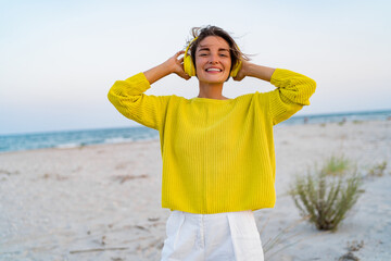 happy smiling woman listening to music in colorful yellow headphones on beach in summer