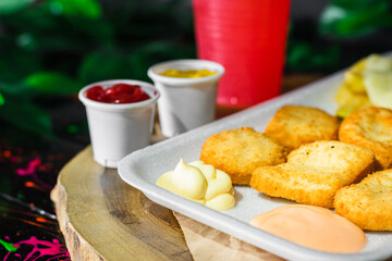 close-up detail of a plate of chicken nuggets with potatoes and mozzarella cheese, served on a wooden tray with a glass of red fruit juice next to it, with two small glasses of sauce.