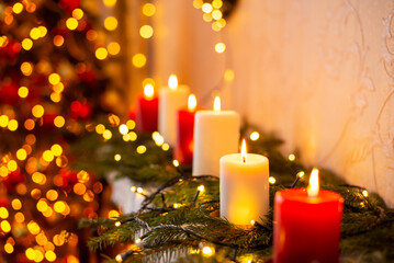 Magical vibes, yellow warm colours, festive interior. Burning white and red candles, shiny lights of garland and blurred christmas tree on the background