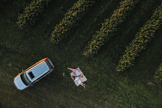 Aerial top-down shot of couple enjoying picnic on road trip in the beautiful fields, man and woman lying on a blanket near the SUV car at sunset