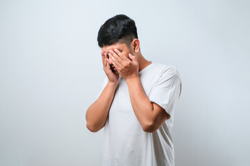 Asian young man wearing white shirt with sad expression covering face with hands while crying.