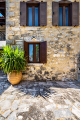 Stone cottage facade with 3 windows and open brown shutters, plant pot besides. Greece, Crete. 