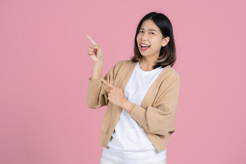 Smiling young asian woman standing pointing index fingers aside up on mock up copy space isolated on pink background studio portrait