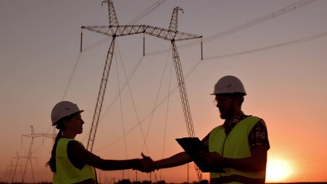 Two electrical engineers meet and shake hands against the backdrop of high-voltage electrical tower at sunset. Meeting of workers near power grids. Teamwork, signing contract, businessmen Slow motion