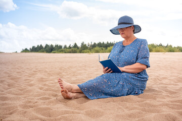 Elderly woman reads a book on the sandy shore.