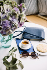 Delicious fresh morning espresso coffee, blue notepad and handbag, cookie, sunglasses and purple hydrangea flower in a vase in the white table background, top view, flat lay