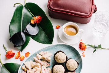 Delicious fresh morning espresso coffee with chocolate and sweets, sunglasses and a red hand bag on the white table, decorated with green leaves and red flowers